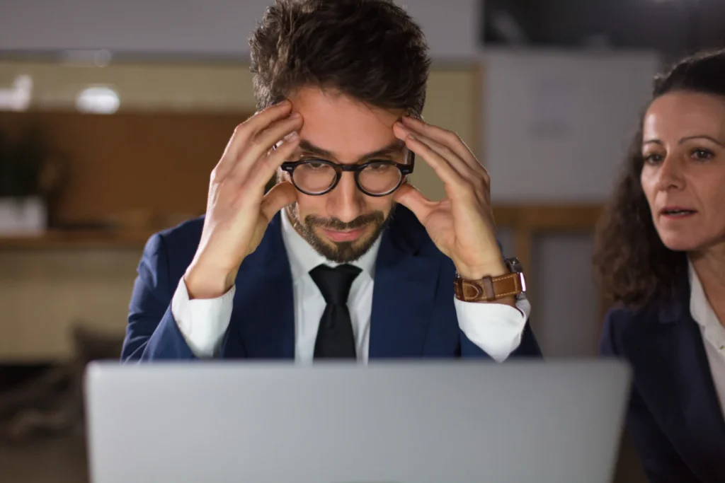 Homem de terno com óculos ajustando a cabeça enquanto tenta resolver problemas na presença digital da sua empresa, ao lado de uma mulher esperando.