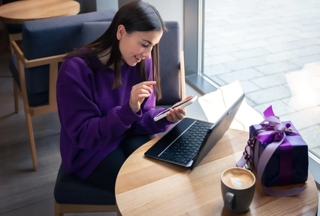 Passo a Passo para Lançar Sua Loja Virtual em Poucos Dias: Mulher sentada em uma mesa usando notebook e celular enquanto organiza detalhes de uma loja virtual, com pacote de presente roxo e caneca ao lado; ambiente interno com janela ao fundo.